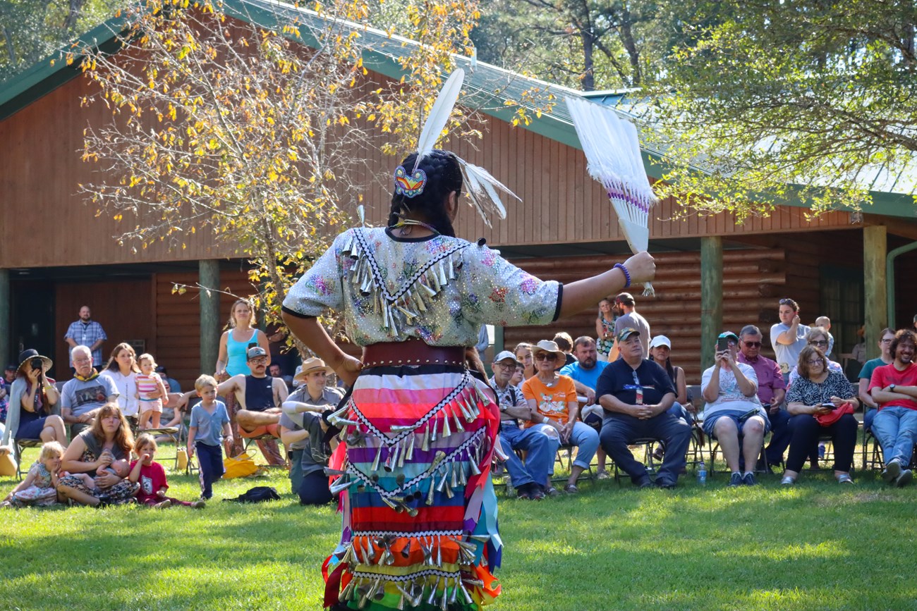 Alabama-Coushatta dancer An Alabama-Coushatta dancer wearing a rainbow-colored dress with shiny bells and white feathers. She is dancing on a grassy lawn in front of a seated audience with the visitor center in the background.