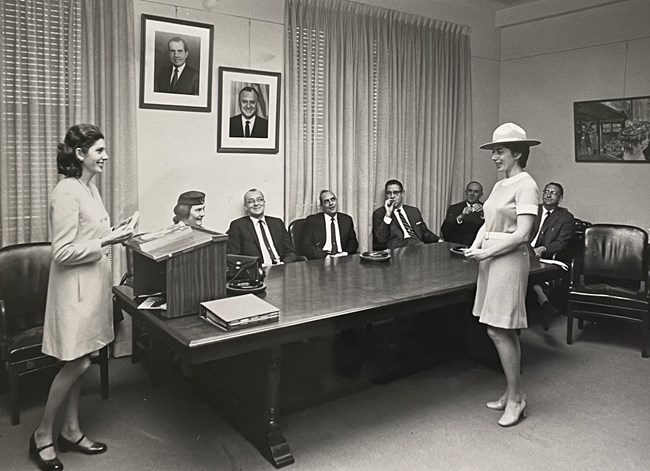 Two women stand as a woman and several men sit around the table
