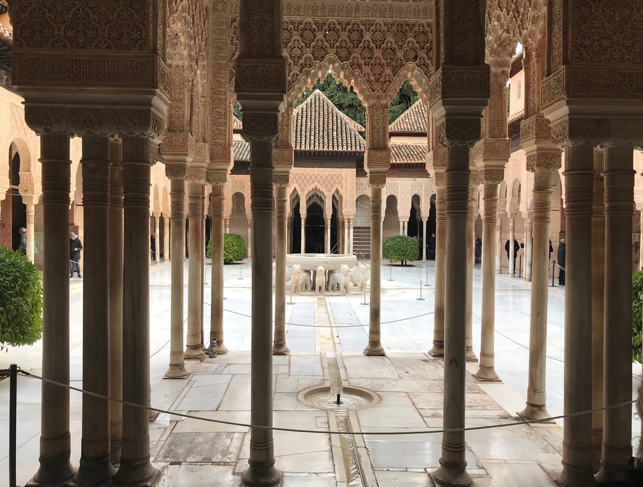 View between several ornate columns leading to a courtyard with a concrete fountain surrounded by decorative animals.