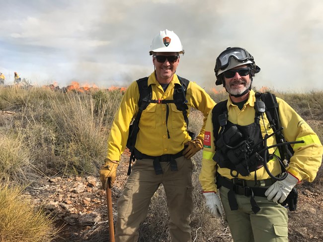 Two wildland firefighters wearing all of their personal protective equipment stand in a rocky area of a field with flames from a prescribed burn in the background. Two other firefighters are in the upper left background.