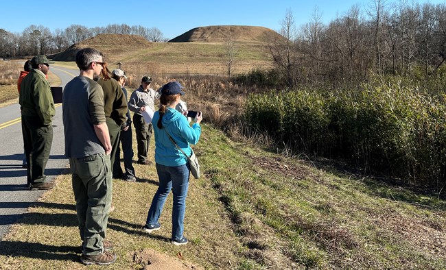 People standing on a road with a mound in the background, looking at vegetation.