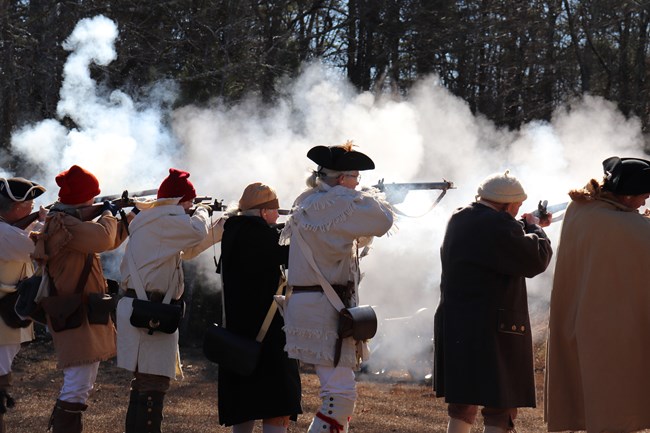 Several reenactors in period clothing fire muskets with large cloud of smoke.