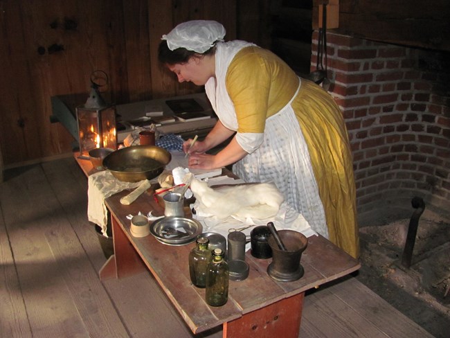 A woman in 18th C clothing leans over a table with various bowls and a lantern.