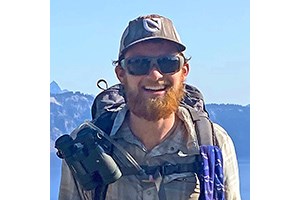 Will Babb Headshot of will in hiking gear, wearing a baseball cap, backpack, sunglasses, and a beard.