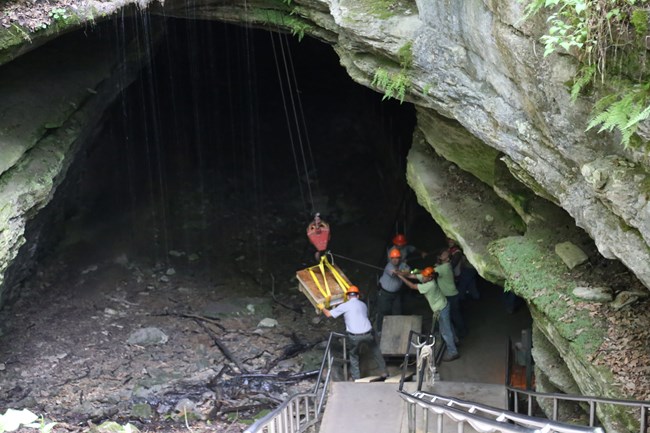 A crew of people wearing hard hats lower a large stone into the cave using a crane.