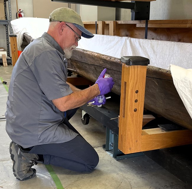 Man kneels next to wooden canoe, preparing to treat it
