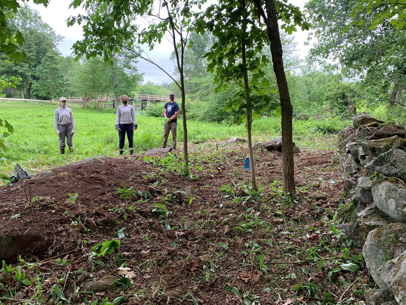 Three workers, two women and a man, stand apart from a stone wall and from each other. They are wearing cloth face masks. Behind them is a grassy field bordered by forest. A bridge is in the distance.