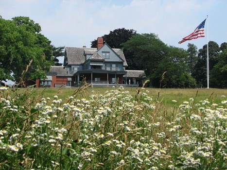 A large light blue house sits in the distance of a field.  A flag pole with an American flag is seen on the right.