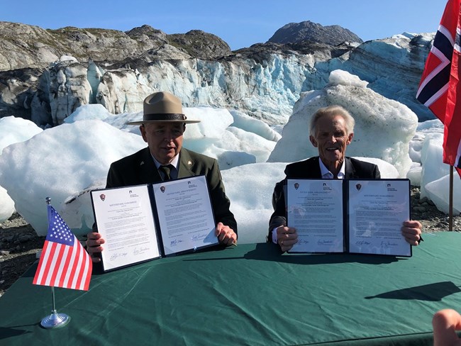 two people holding signed papers, with ice and snow in the back, and half of the norway flag can be seen on one side