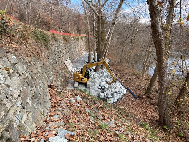 A long stone wall holds up a pathway on the left, while construction equipment builds a buttress of stone at the bottom