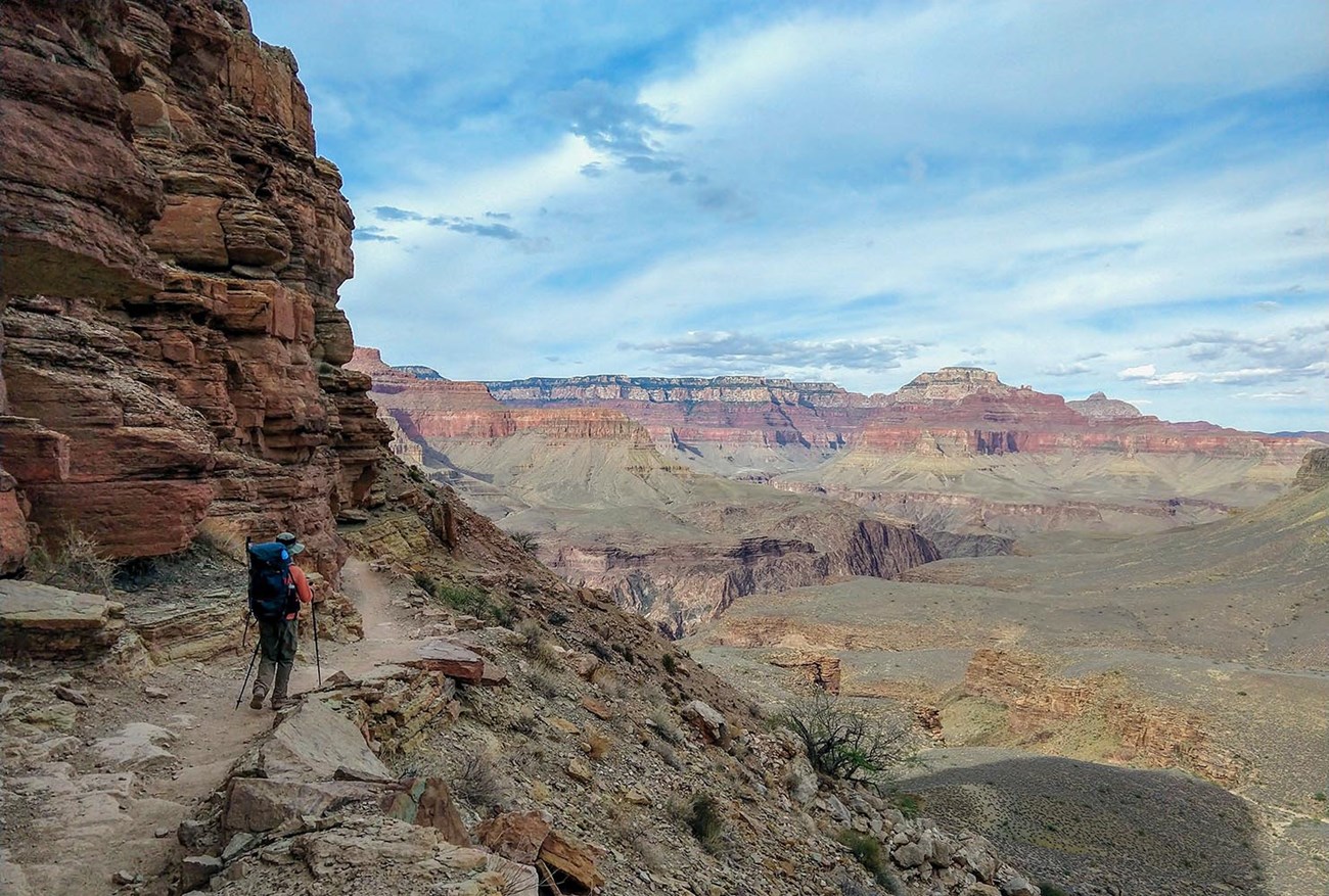 IMG_20210601_172017585 South Kaibab Trail. NPS Photo/K. Hensel A person with a backpack and trekking poles is walking on an unpaved trail built into the side of a cliff. In the distance colorful peaks rise from the floor of a canyon.