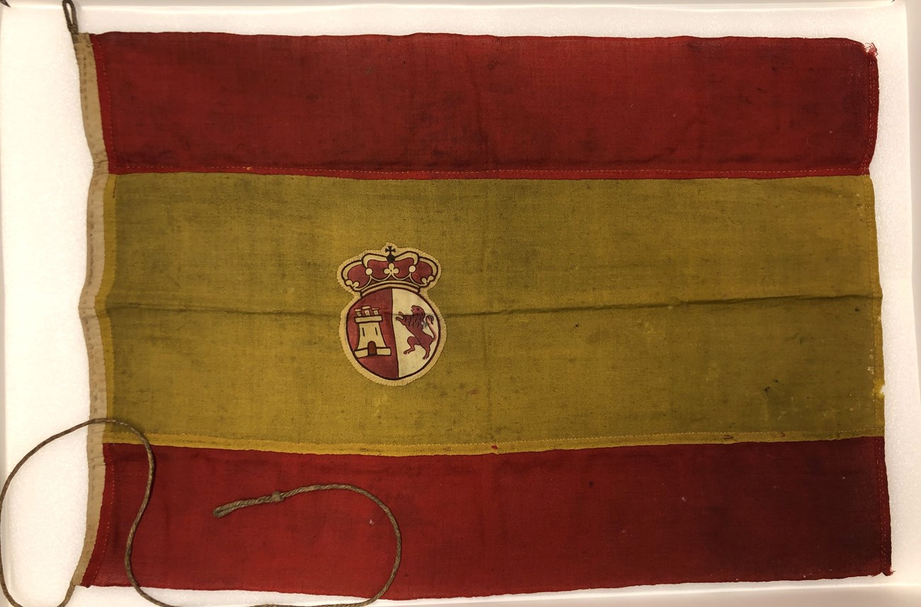 Spanish Flag FOVA A photo of a Spanish flag, with two red stripes and one yellow stripe. The seal of Spain is located in the yellow stripe. The flag is worn.