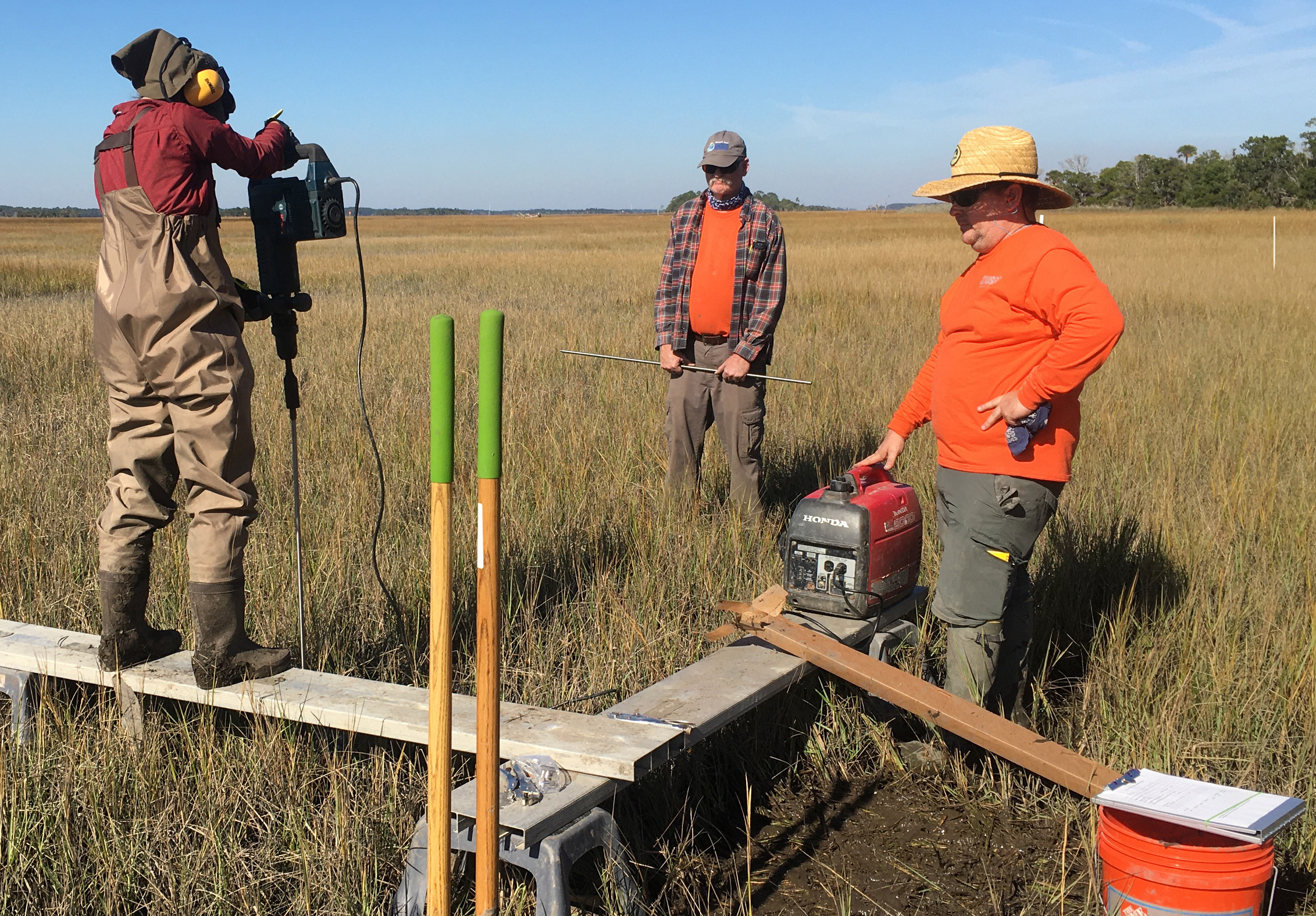 Woman using jackhammer on a platform with two other people standing by in a marsh.