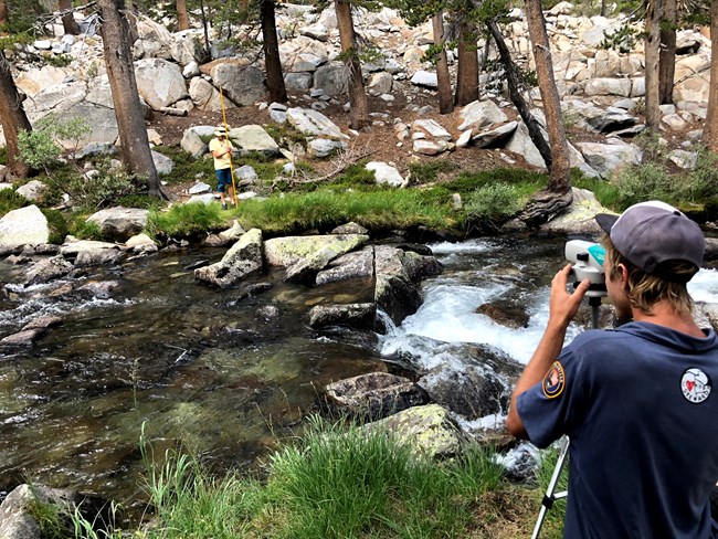 Scientist looks through instrument on a tripod to another person standing on opposite side of a river, surveying the channedl.