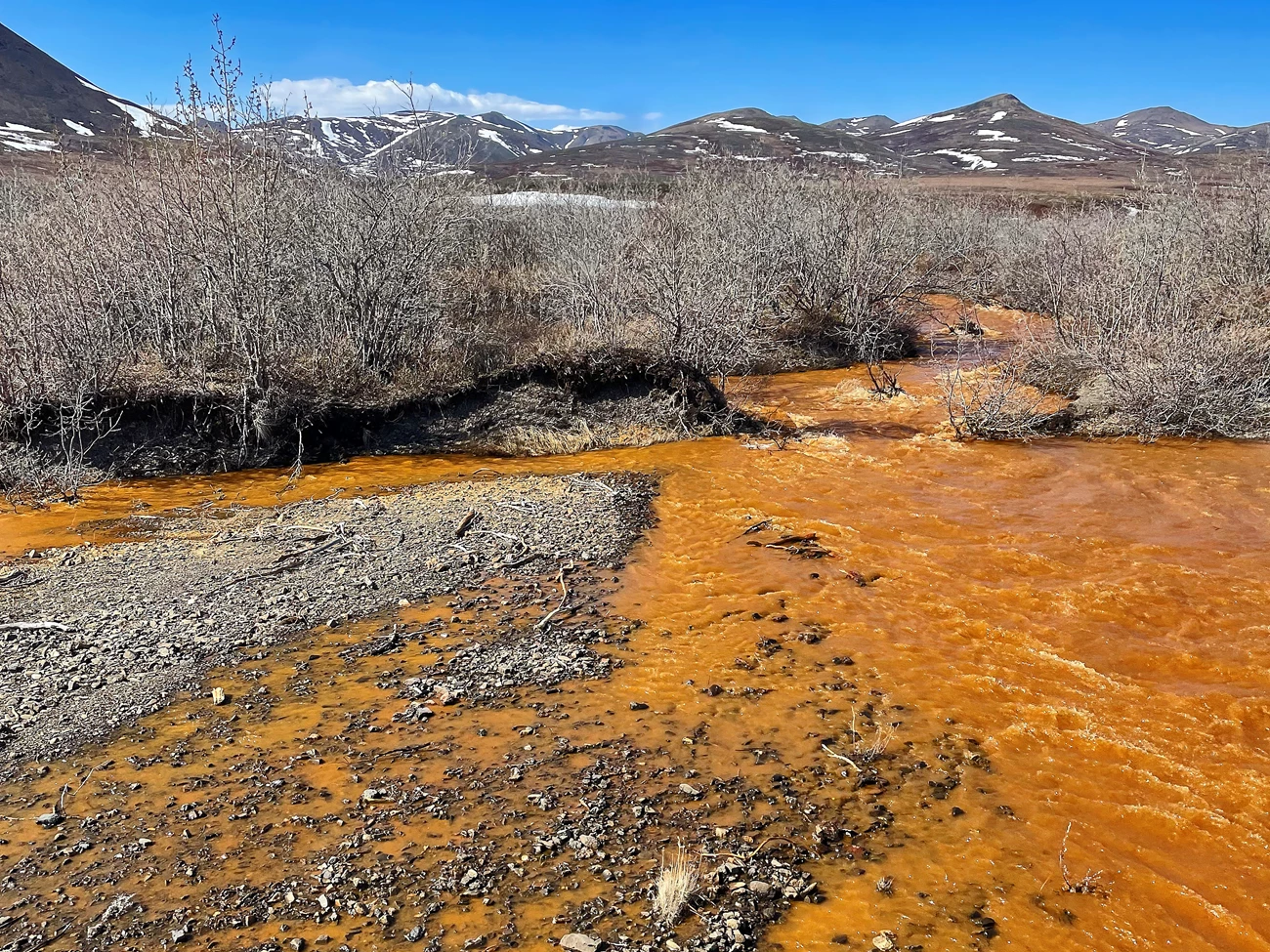 Orange tributary of the Kugororuk River Rushing stream where all of the water is bright orange. Mountains covered in patches of snow line the horizon.