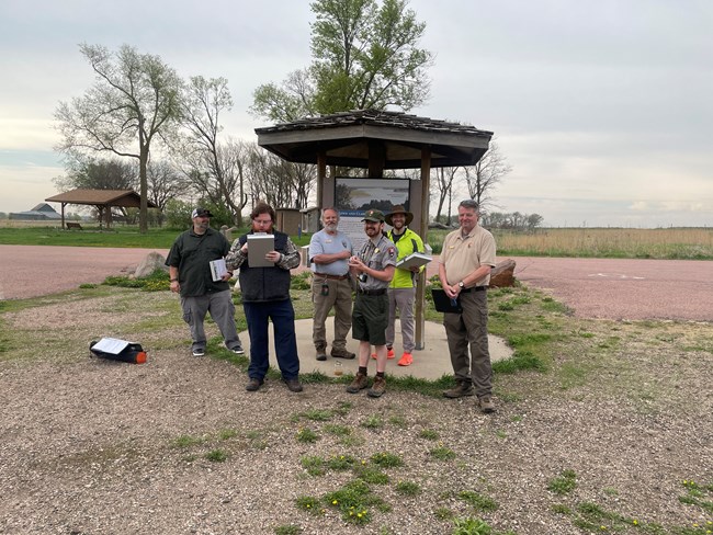 Six people stand in front of an information sign. They are holding notebooks and pens.