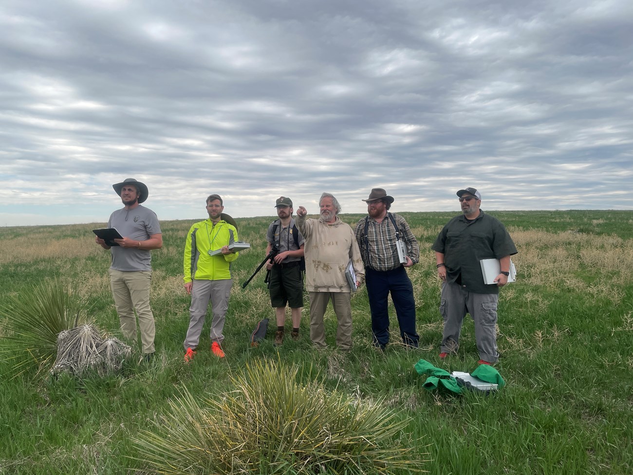 Five people standing in a field carrying surveying equipment and notebooks. One is pointing off into the distance.