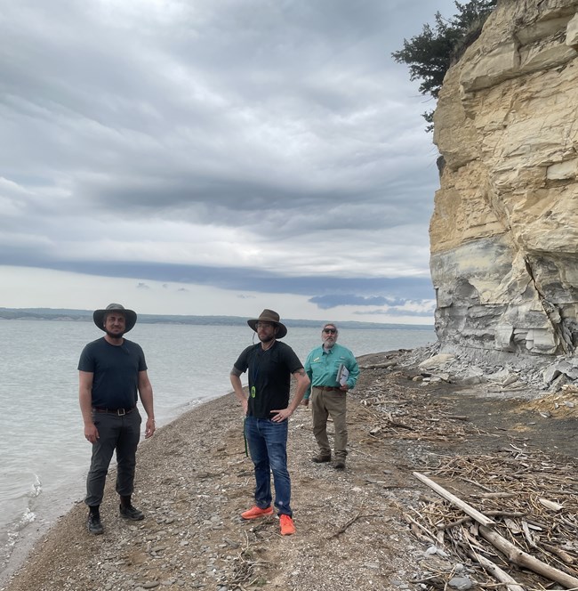 Three people stand on a rocky lakeshore. To the right is a tall, vegetated cliff face.