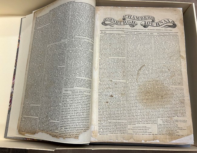 An open book shows pages of historic Scottish newspapers. The newspaper pages are yellowing and have pieces missing on the edges. The newspapers are mounted on white paper for their preservation