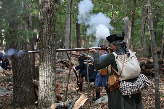 A colonial soldier with knapsack and blanket fires his musket in the woods.