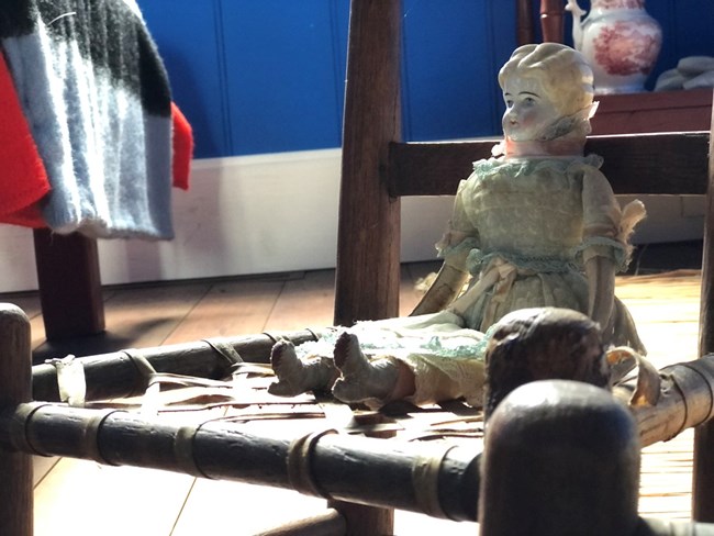 A porcelain doll wearing a dress sitting on a child sized chair in a room lit by natural light.