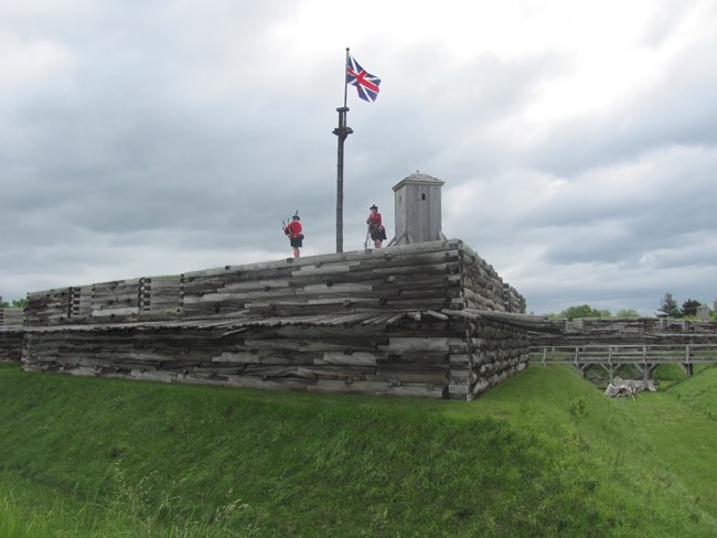 Two men in kilts walk across the fort wall under a British flag.