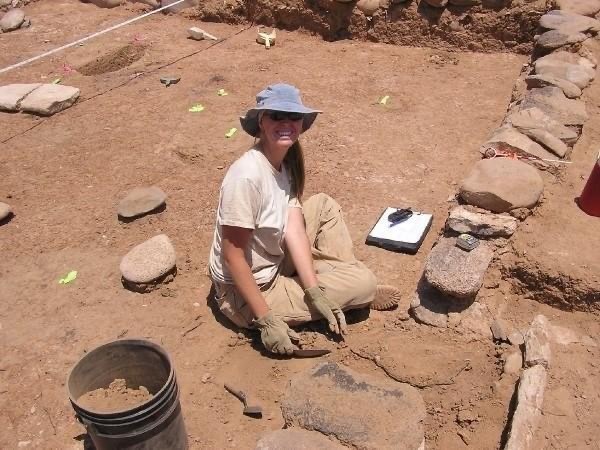 Archaeologist sitting on ground with tools and notebook at a field site.