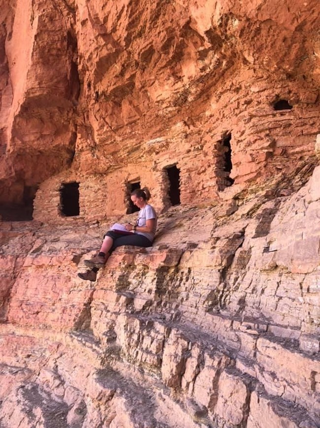 Archaeologist sitting near cliff dwelling taking field notes.