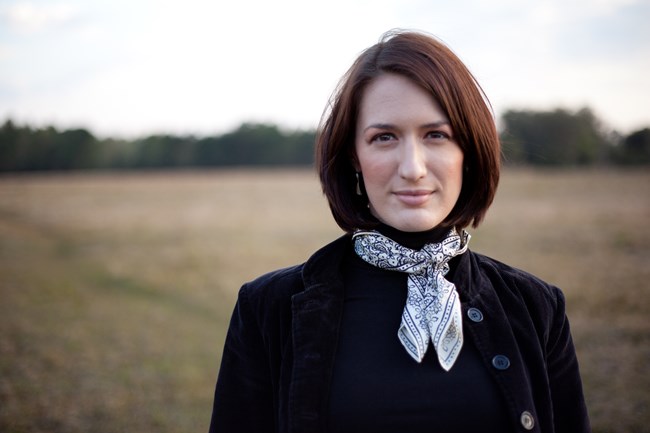 Woman with red hair wearing a dark velvet jacket and a white patterned scarf around her neck stands outdoors at the edge of a field