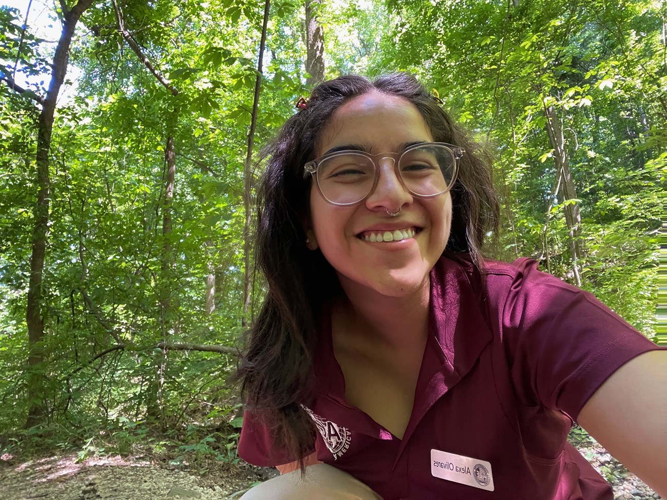 Intern Alexa Olivares at GWMP- Turkey Run Park. They are taking a selfie in nature wearing an ACE shirt, glasses, and a hair clip.
