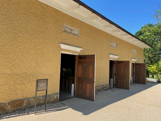 Side view of the South Slave Quarters at Arlington House. There are three wooden doors on a yellow building. There is a sign saying "South Slave Quarters".