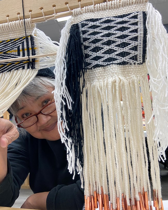 Tlingit_Weaving-IMG_4018 Woman smiles at camera while holding a woven item to the side.