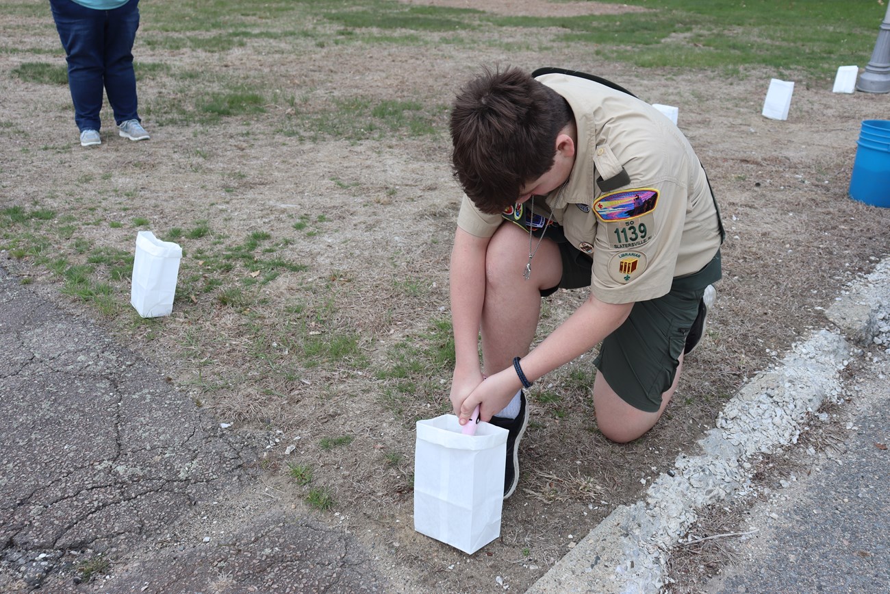 a boy in a boy scout uniform kneels on the grass lighting a candle inside a paper lantern