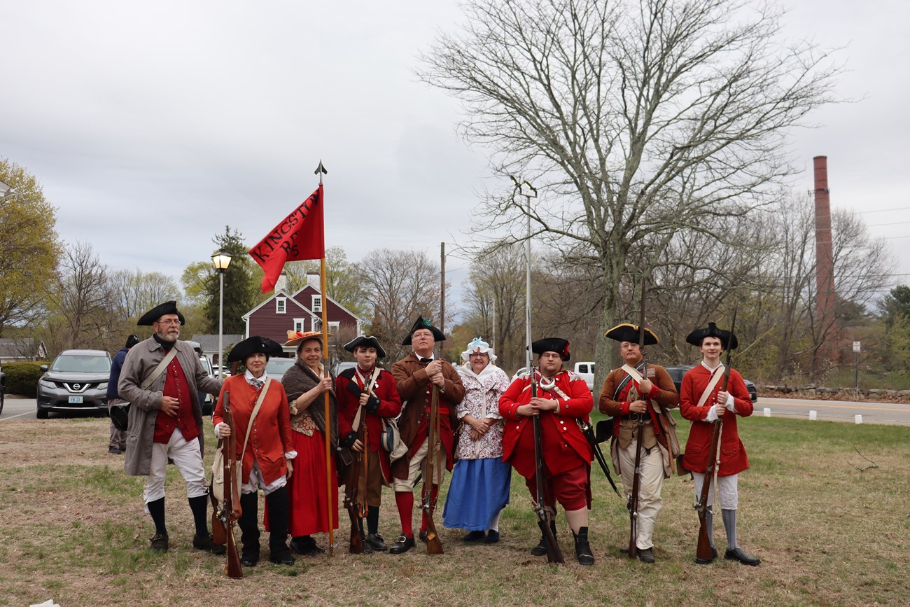 A group of historic reenactors dressed in red, brown, and blue colonial clothing holding historic weaponry