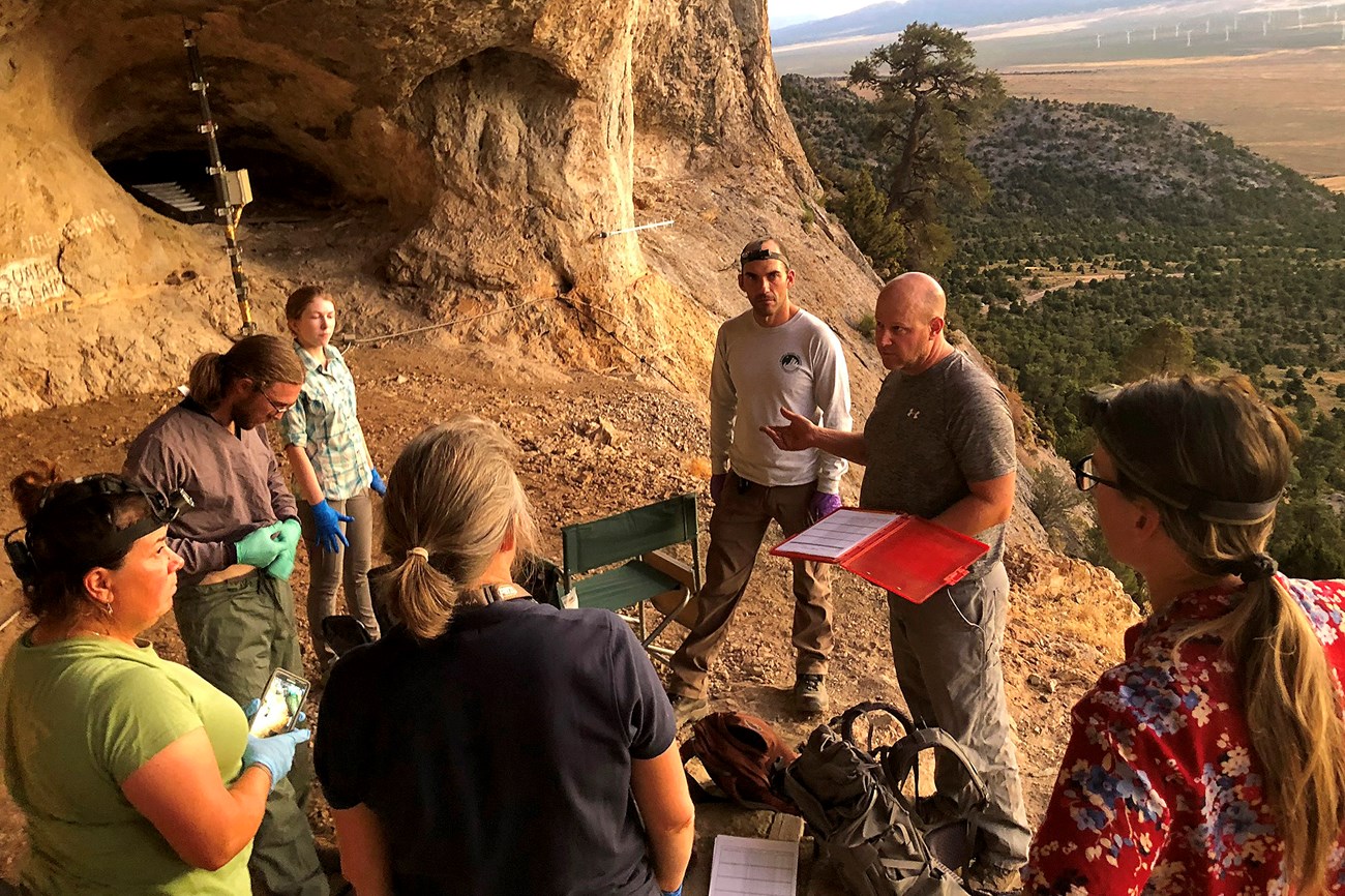 Biologist at the mouth of a cave speaking and gesturing to a half dozen colleagues equipped with rubber gloves, headlamps, and backpacks. Audio recording devices are set up nearby.