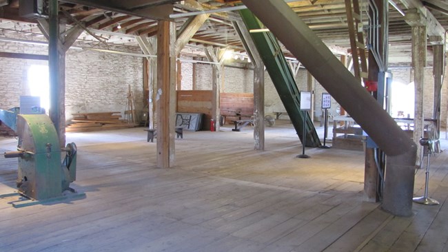 Inside barn with wood floors and limestone walls. Grain grinding machinery, grain chute, and other equipment visible.