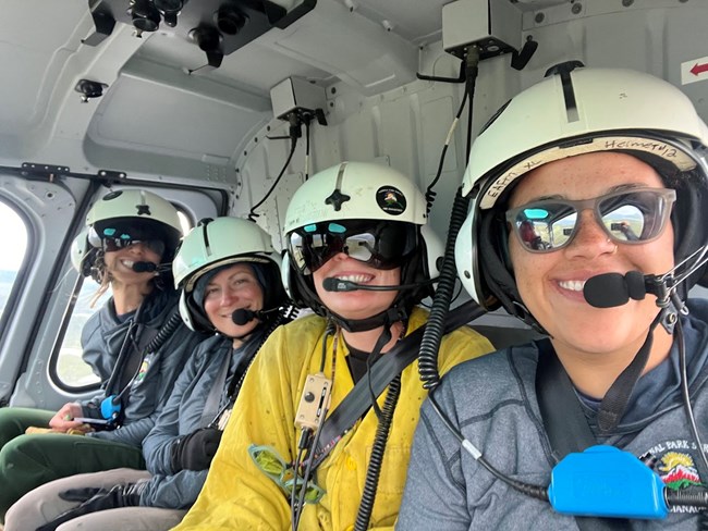 Closeup of four women wearing helmets and smiling at the camera from inside a helicopter.