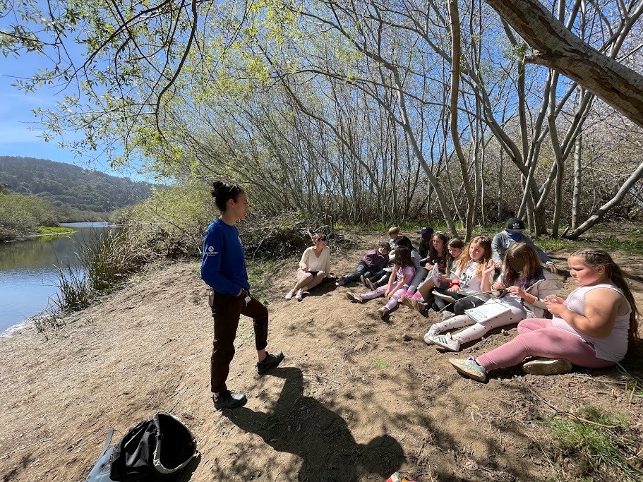 Field trip Corpsmember standing in front of a group of children outside beside a calm waterway.