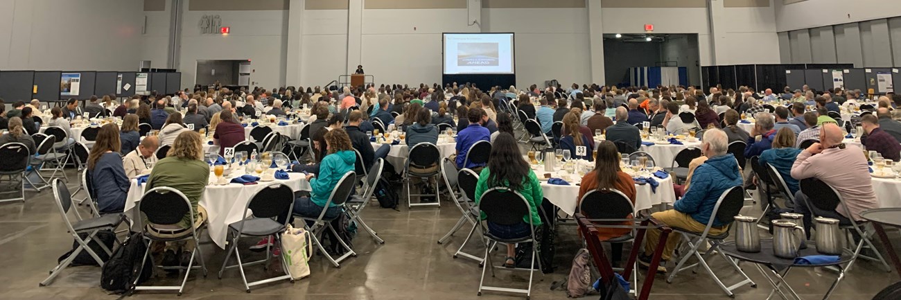 13th National Monitoring Conference Large room full of people sitting at round tables with a podium and large screen in the background.