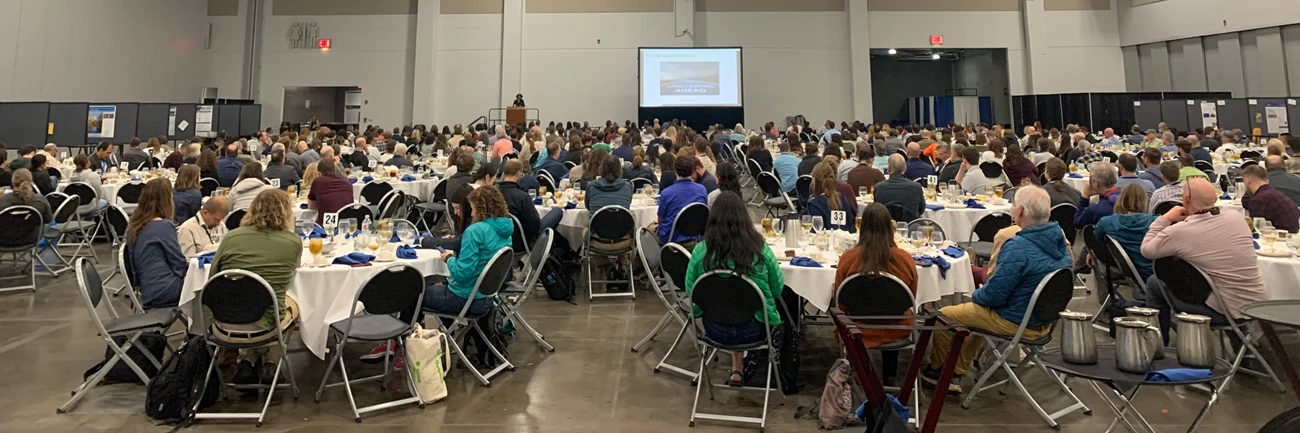 13th National Monitoring Conference Large room full of people sitting at round tables with a podium and large screen in the background.