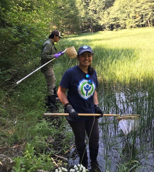 A smiling girl holds a net while standing in the water along the shore of a lake. A second person looks at a net in the background.