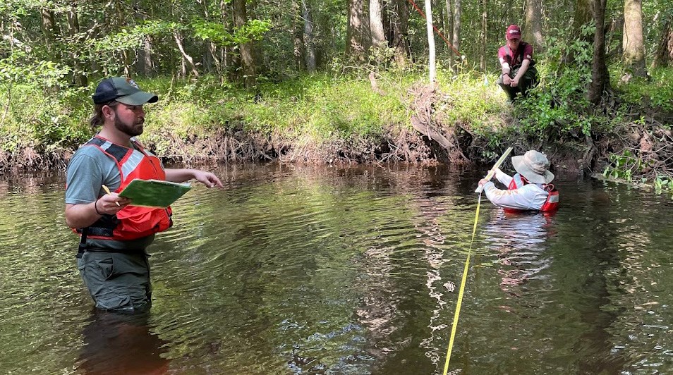 Man standing in stream with clipboard, another man in the water holding a tape by the bank and a woman kneeling on the bank