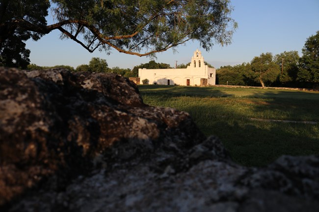 Limestone half-walls in foreground surrounded by green grass. Mission San Juan white limestone church facade in background.