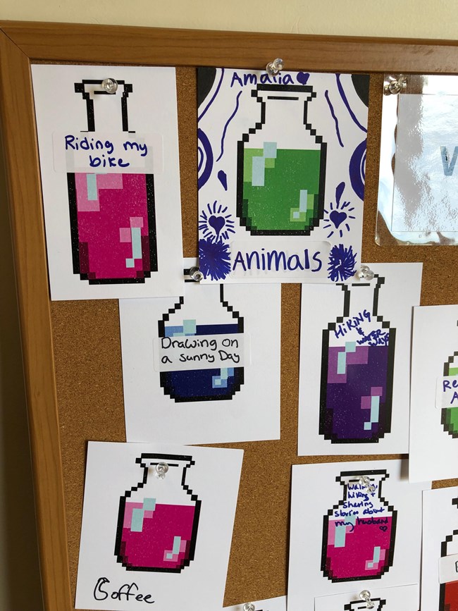 A cork board with colorful bottle illustrations bearing hand written labels.