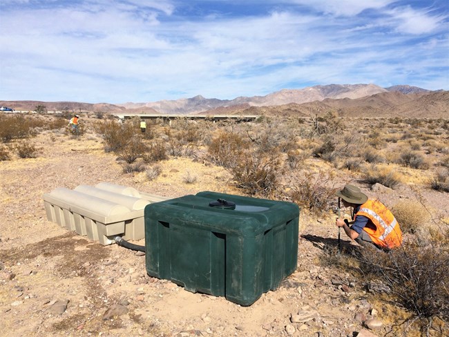 woman in the desert squats next to large plastic crates