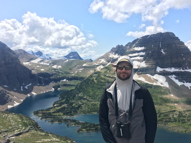 Man in jacket with binoculars, standing in front of rugged mountain peaks and a deep blue lake, Glacier National Park