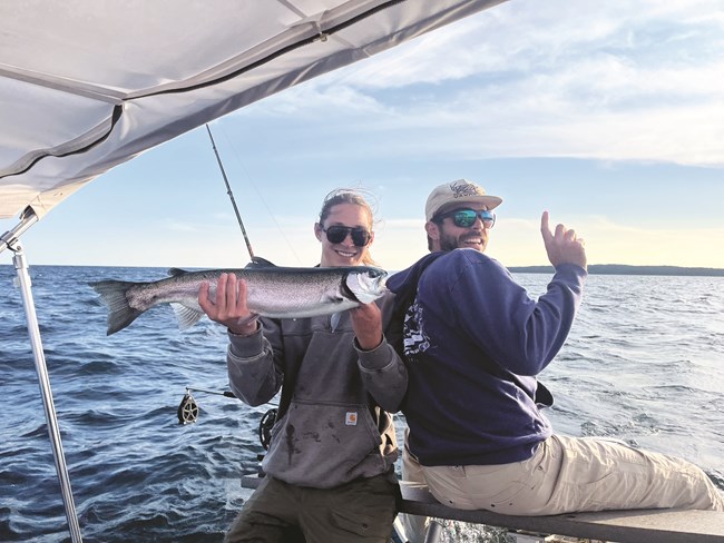 Two people on a boat; one holding a freshly caught lake trout, and the other holding up their index finger and looking at the camera.