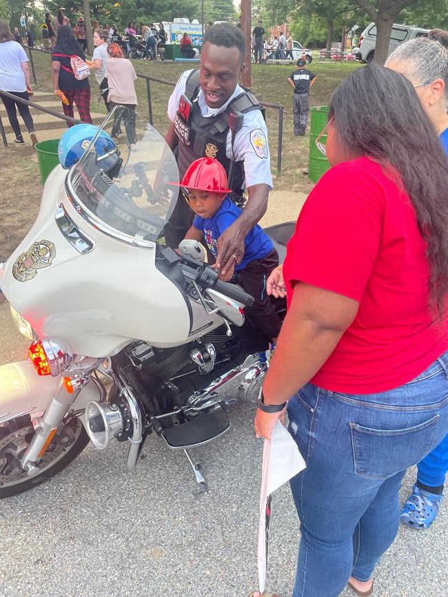 Boy Riding USPP Motorcycle National Night Out A U.S. Park Police Officer in uniform shows a young boy wearing a red firefighter hat who is sitting on a U.S. Park Police motorcycle how the motorcycle works. Two women watch from the side.
