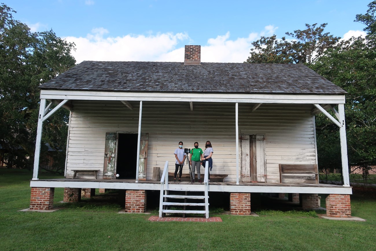 Three women stand on the porch of a tattered white house.