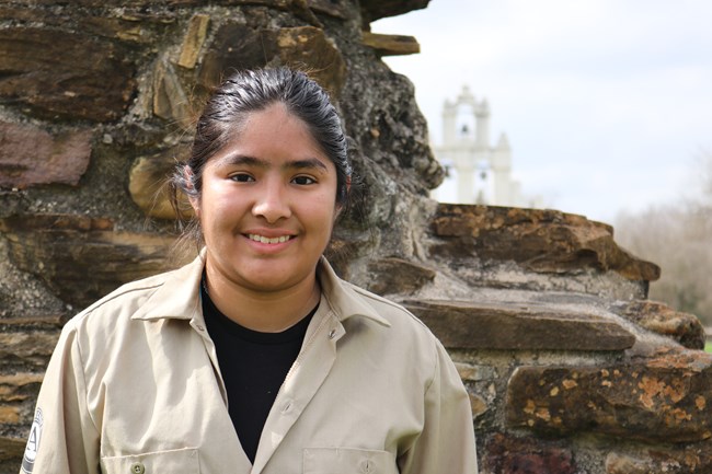 Apprentice Yukary stands in front of a limestone wall with the bright white church facade in the background at Mission San Juan
